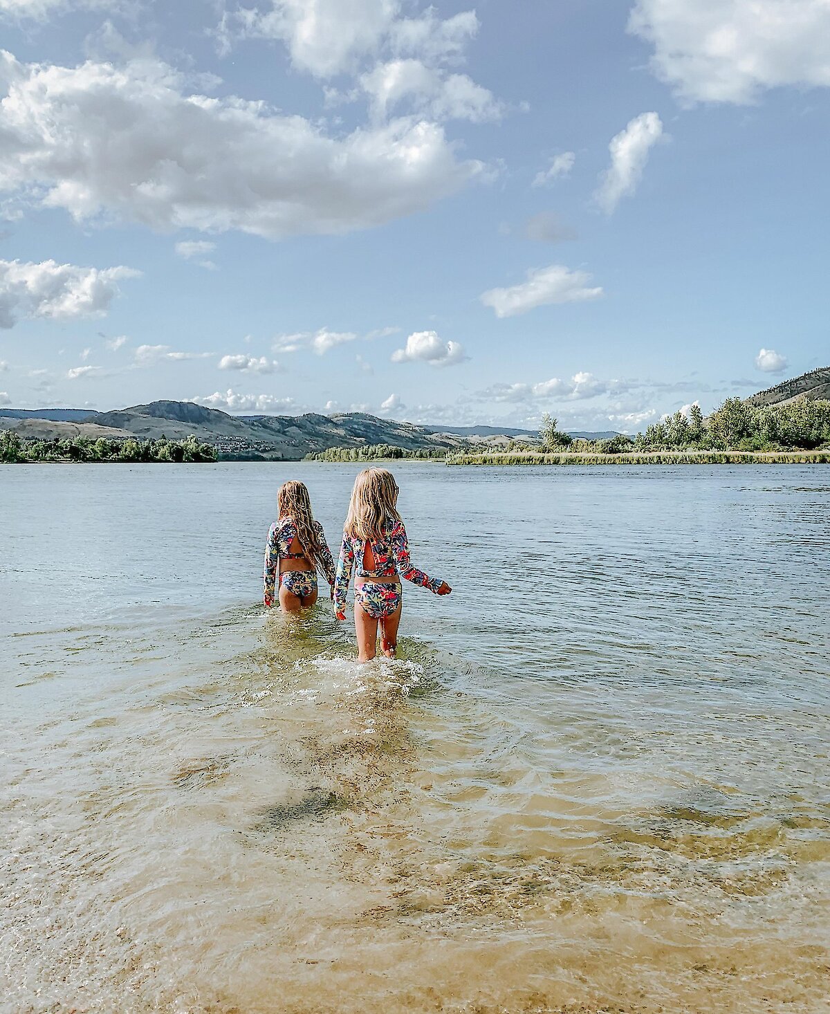 Two young girls wading into a calm river with mountains in the background near Kamloops