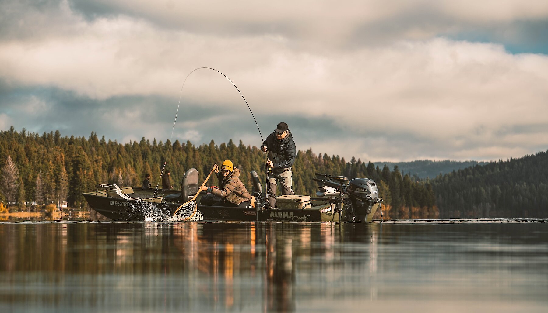 A fisherman reels in a catch as another angler prepares to net the fish, with autumn colours lining the shore behind them.