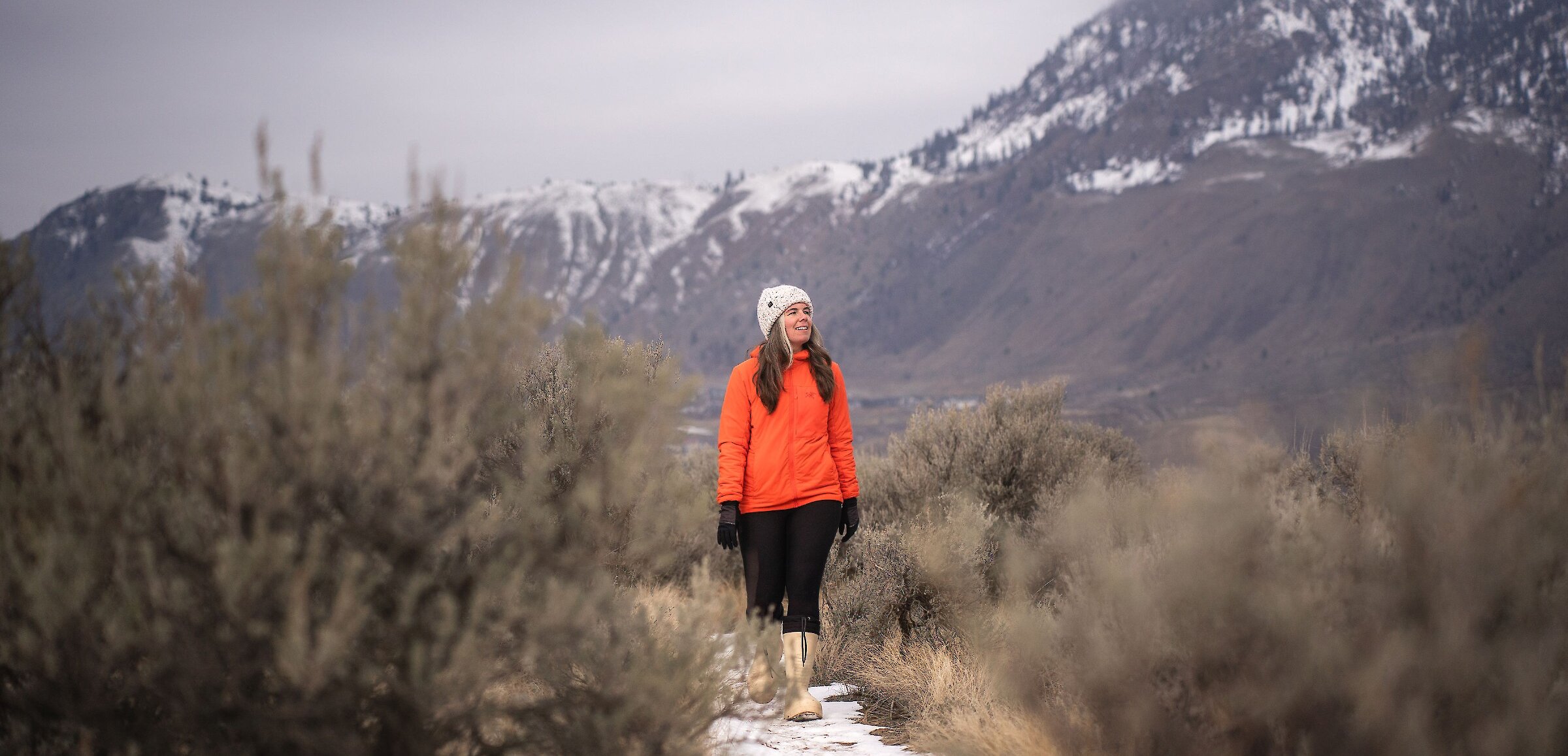A woman walking along the grass ridge with a snow-covered mountain in the background at Valleyview Nature Park in Kamloops, BC.