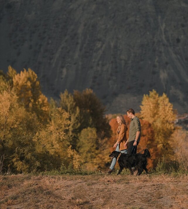 A couple walking their dog along a trail with fall foilage surrounding them.