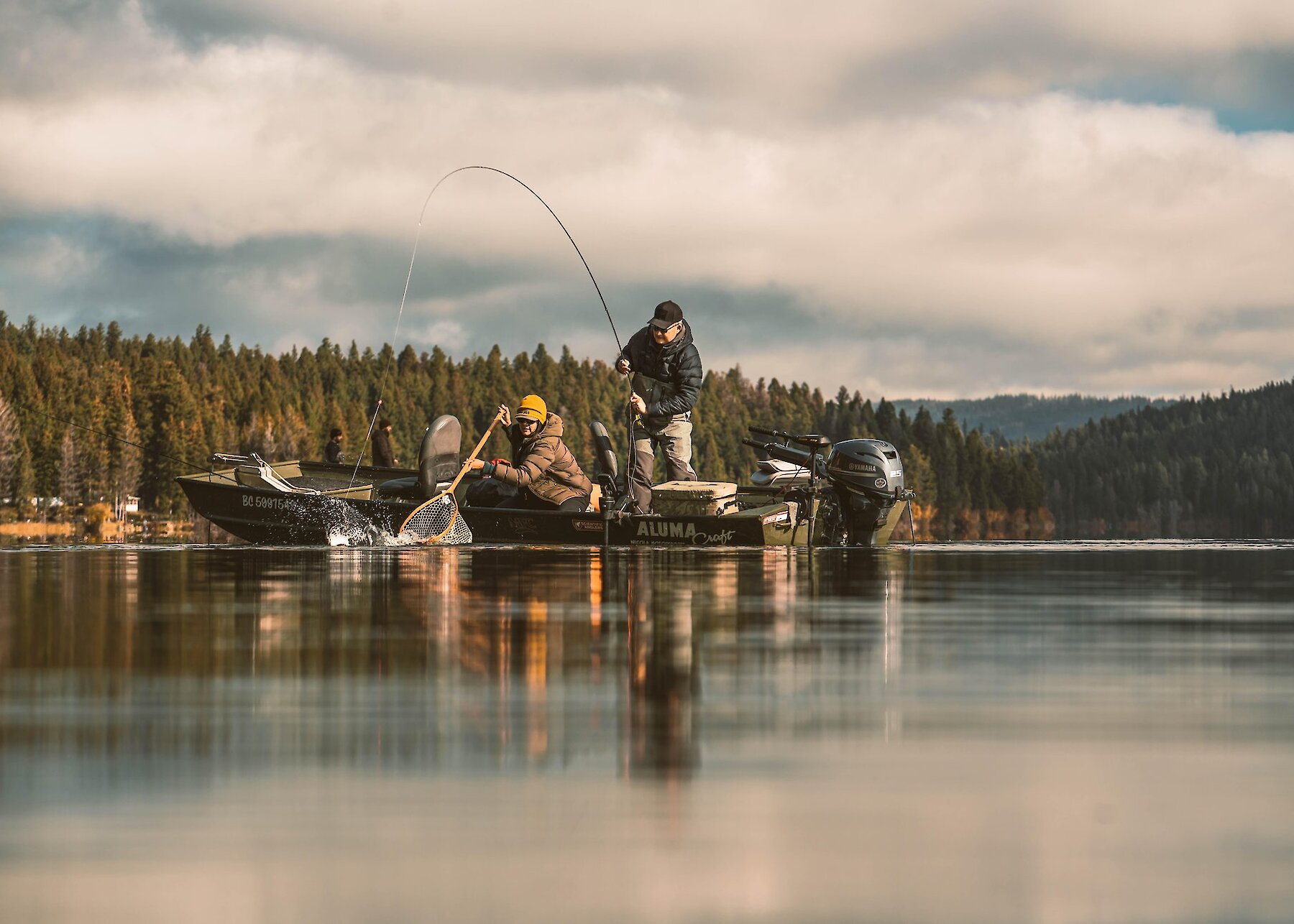 Two anglers in a boat with one reeling in a fish and the other netting the fish as it approaches the boat.