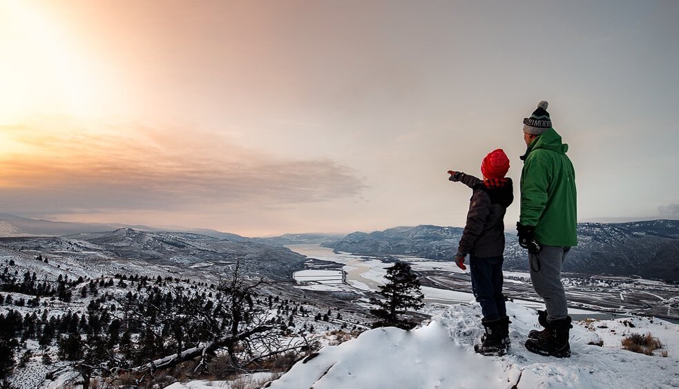 An adult and child stand on top of a snowy hill near Kamloops BC. The child points into the distance towards a pink and orange sunset.