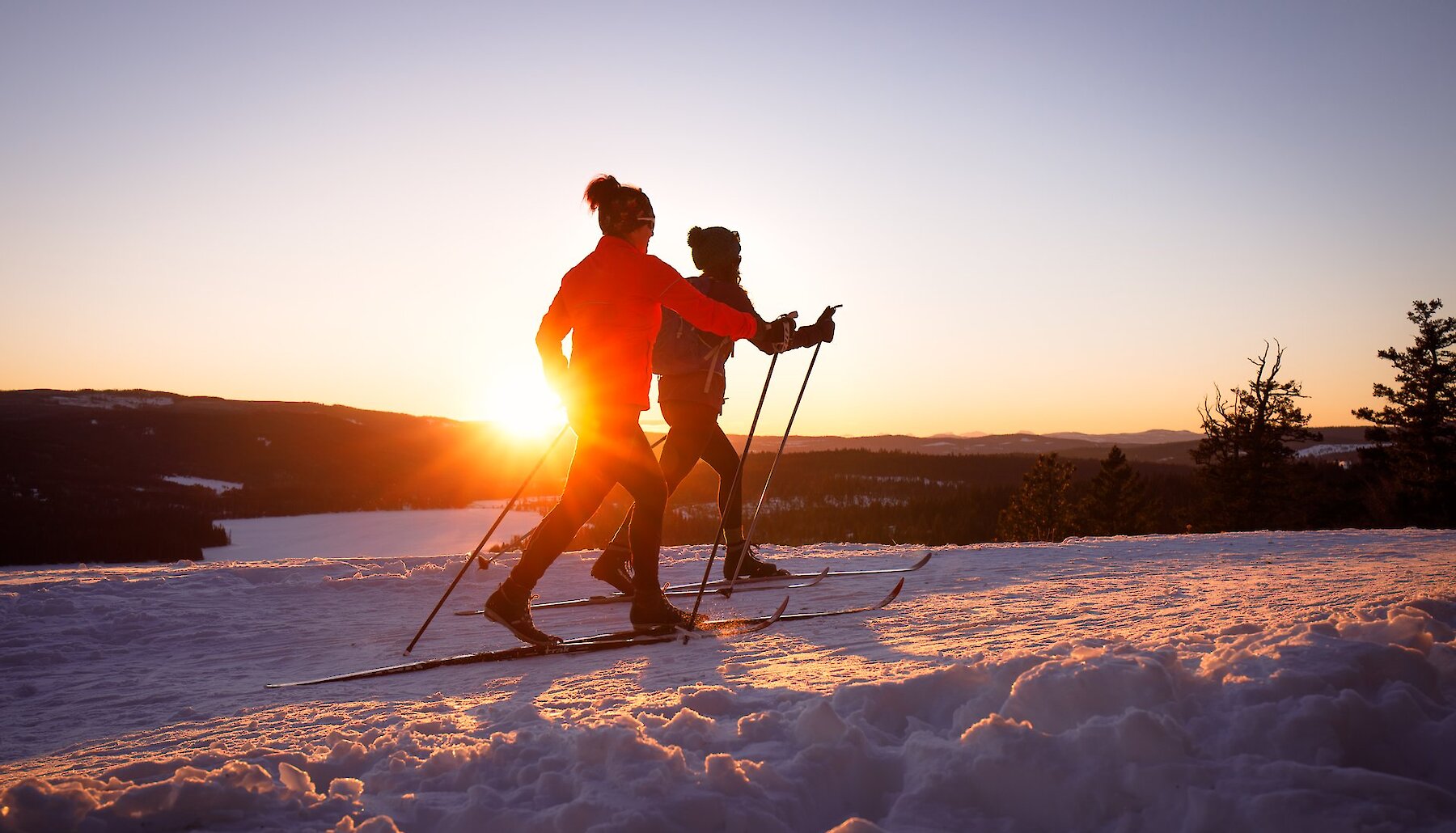 Two people on cross-county skis glide past a frozen lake at sunset, near Kamloops BC.