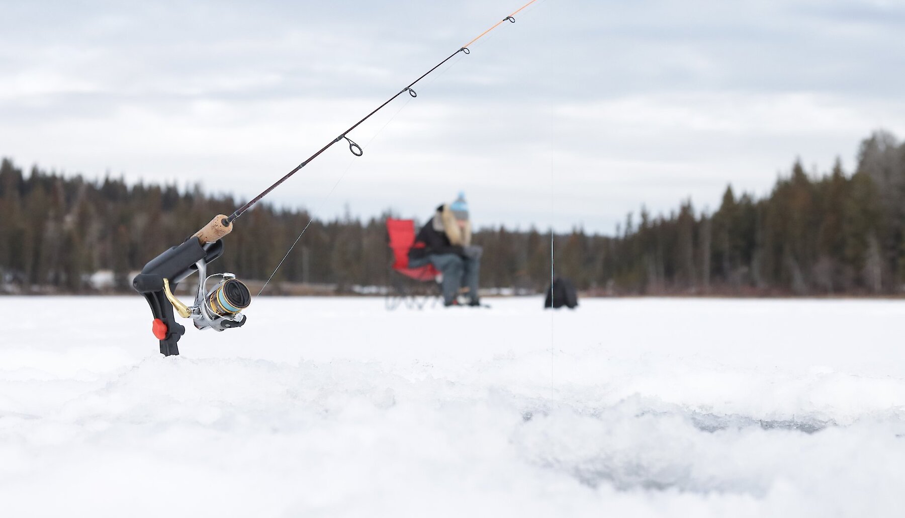 A fishing rod is set up in the snow, the line passing into a hole in the ice beneath it at a lake near Kamloops BC. In the distance, a person sits on a red camp chair, holding a fishing rod with a line into another hole in the ice.