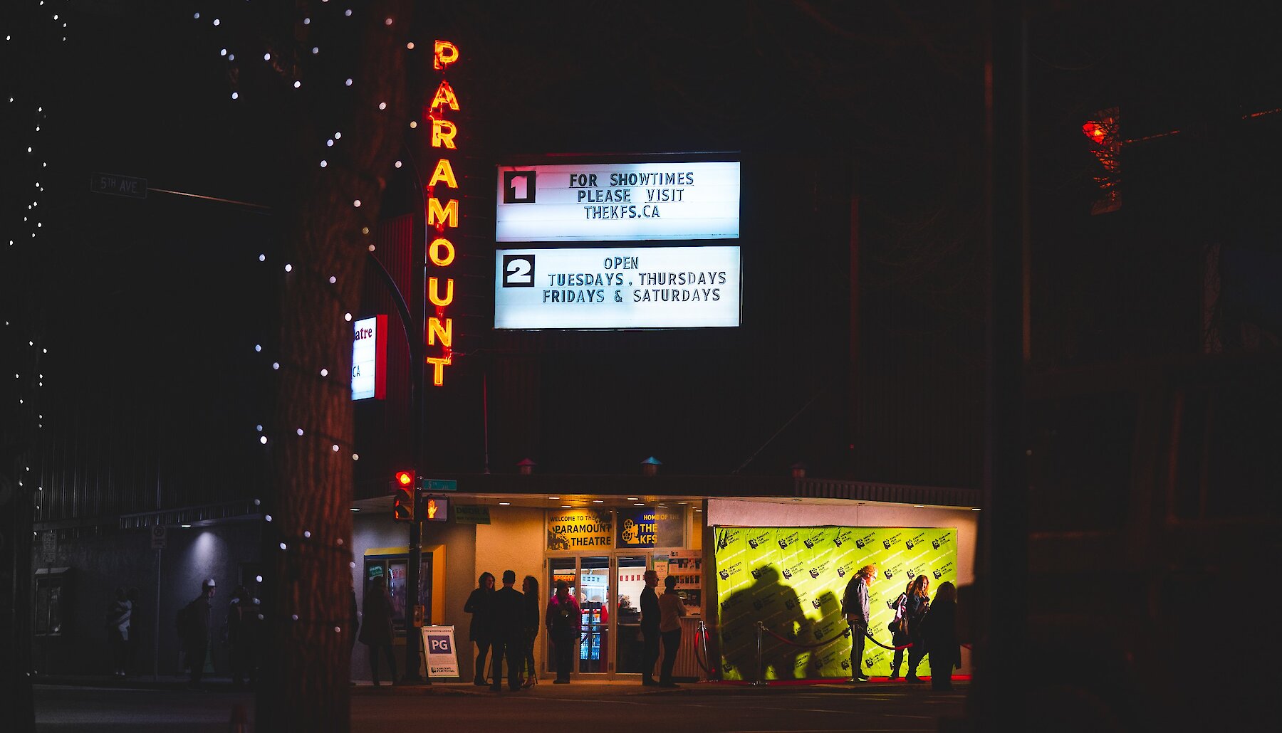 Illuminated Paramount Theater sign in Kamloops,BC at night, with a crowd gathered outside. The marquee displays showtimes and open days.