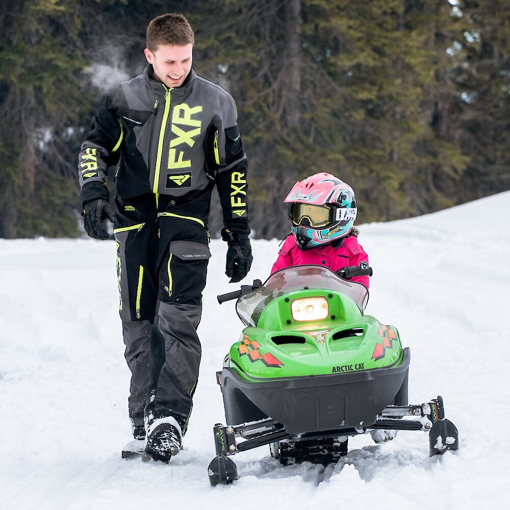 Man dressed in snowmobiling gear walking beside a child riding a child-sized snowmobile near Kamloops, BC.