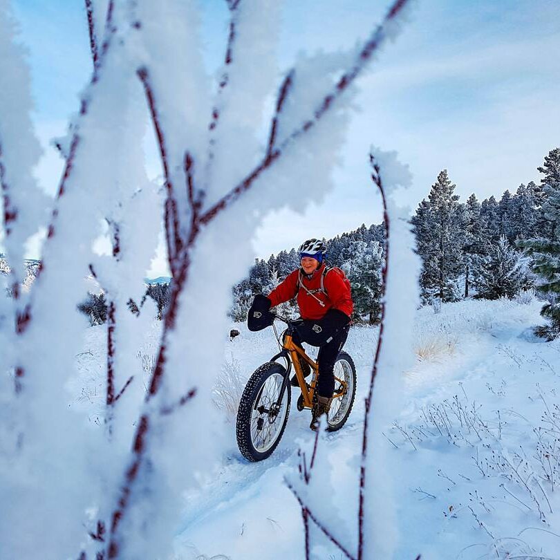 A man rides a snow bike through a winter landscape at Kenna Cartwright Park in Kamloops BC. He is framed by frosty branches in the foreground.