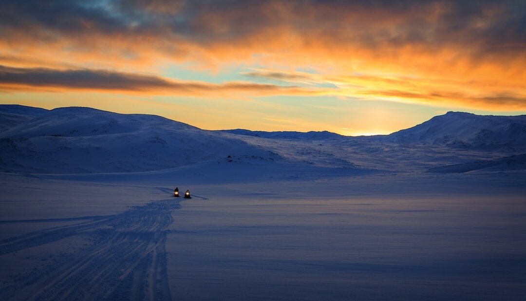 Two snowmobilers traverse an open snowy plateau near Kamloops BC, with hills in the distance and a radiant sunset sky above.
