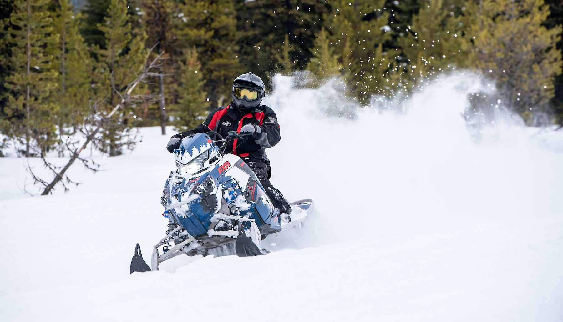 A snowmobiler in a red and black jacket and yellow goggles carves through deep powder, kicking up a spray of snow in a forest near Kamloops BC.