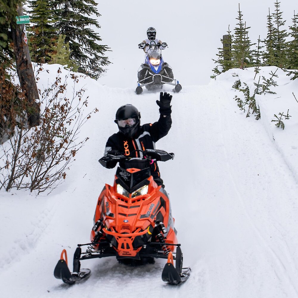 A snowmobilers waves with another rider behind him on a snowy trail surrounded by treees near Kamloops, BC.