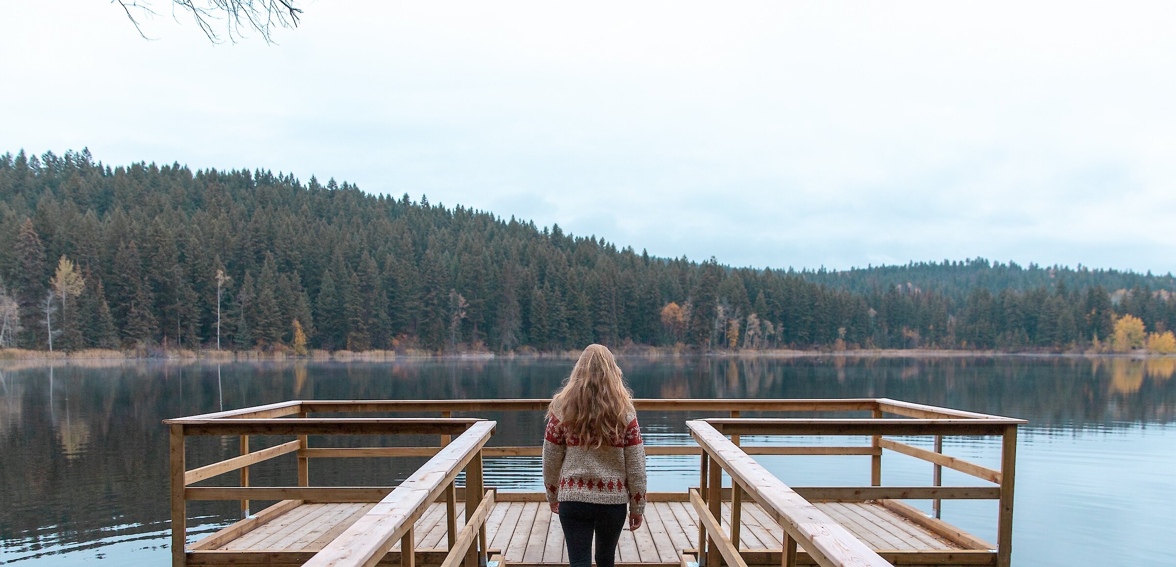 A woman walking down a dock to Isobel Lake with the fall foliage around the shoreline in Kamloops, BC.