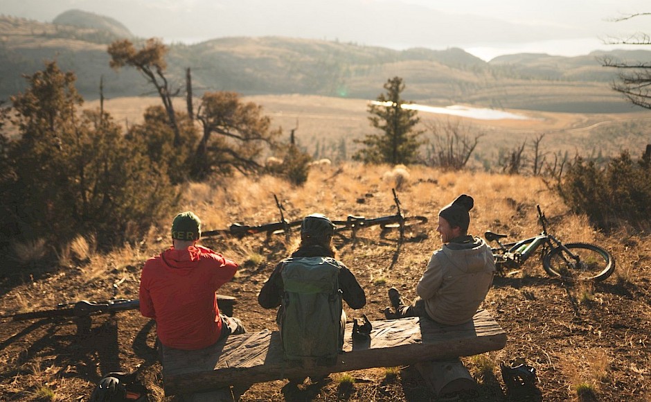 Three people sitting on a bench with mountain bikes in a scenic landscape.