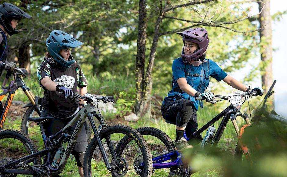 Three mountain bikers, wearing helmets, with their bikes on a trail through a forest.