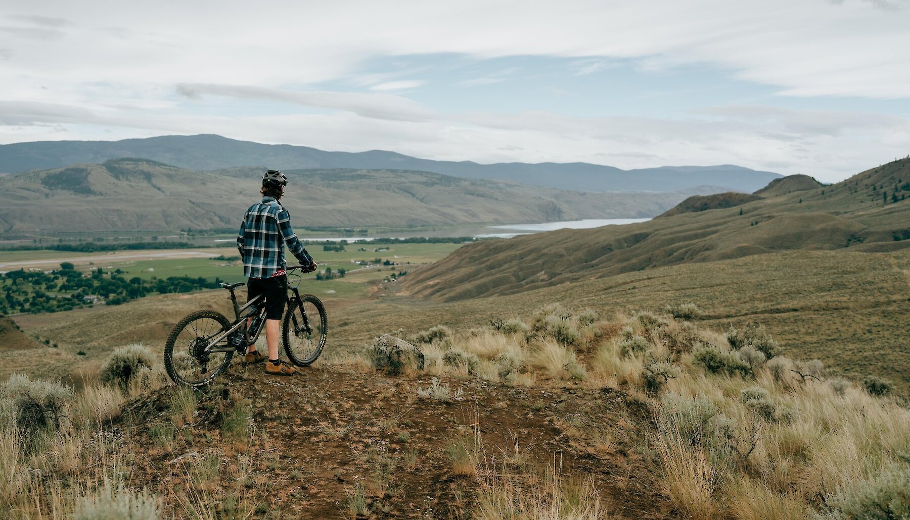 A rider waits on their bike at the top of a ridge, looking out over the grasslands in Lac du Bois, near Kamloops BC