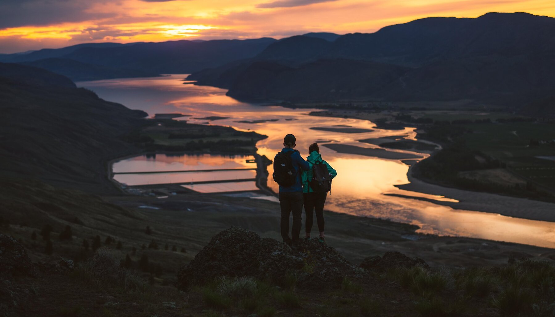 Two hikers look out over the Thompson River Valley near Kamloops BC. A colourful sunset is reflected in the water.