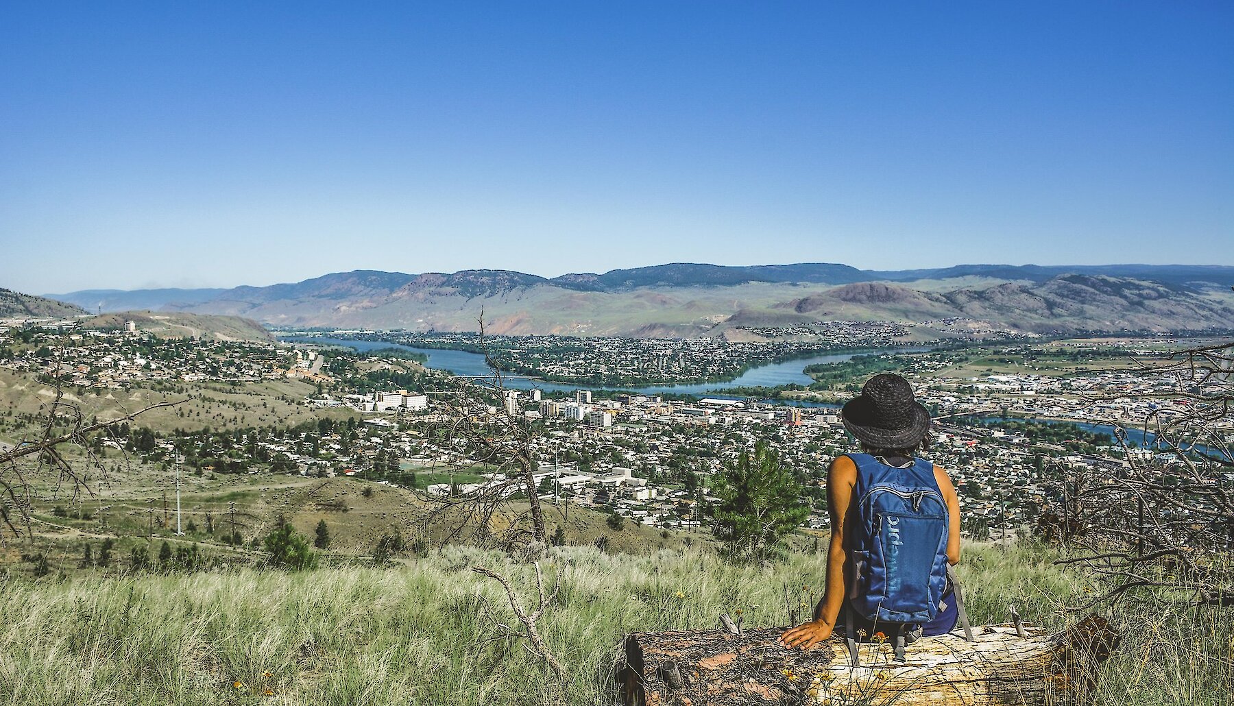 A hiker rests on a log, looking out over the city of Kamloops BC, from a grassy hill, under a bright blue sky.