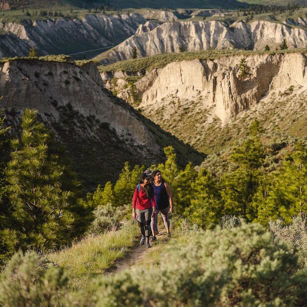 Two hikers ascend a trail through sagebrush and fir trees in a valley near Kamloops BC. Hoodoos and sandstone bluffs are visible in the background