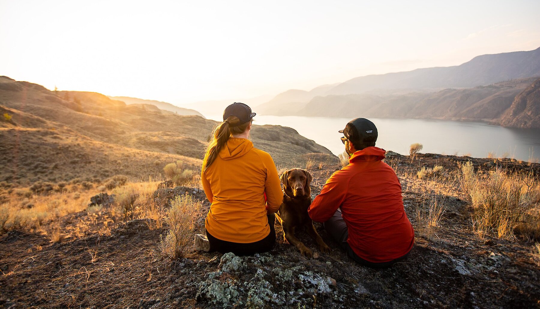 Two hikers rest with their dog on a ridge overlooking Kamloops Lake as the sun sets in the distance.