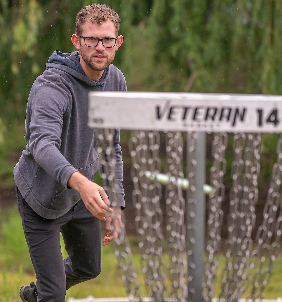 Disc golfer throws a disc towards a metal course basket in a green park setting, surrounded by trees.