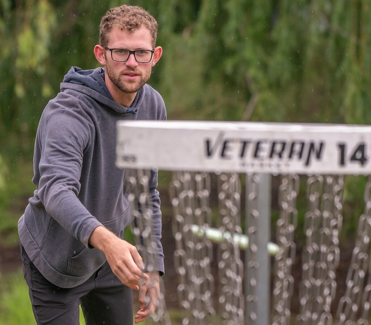 Disc golfer throws a disc towards a metal course basket in a green park setting, surrounded by trees.
