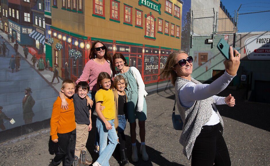 A group of visitors taking a selfie in front of the Wheel in the Sky Back Alley Mural in downtown Kamloops, BC.