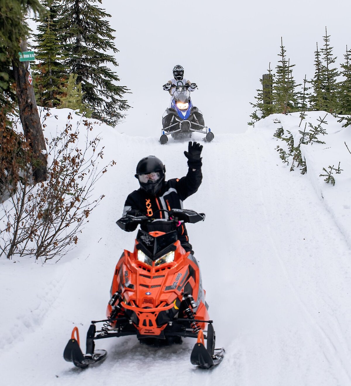 Snowmobile riders on their sleds with Kamloops Snowmobile Adventures and giving the "hang loose" sign with snowy forest in the background.