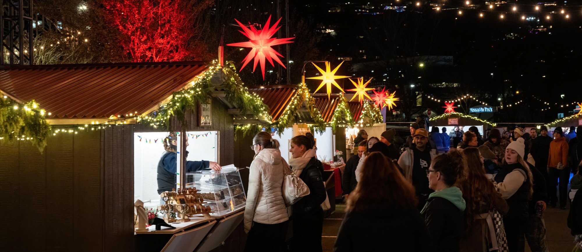 Crowds of shoppers browse the lit wooden chalets at the Kamloops Christmas Market at night, under glowing star lanterns and strings of lights at Riverside Park.