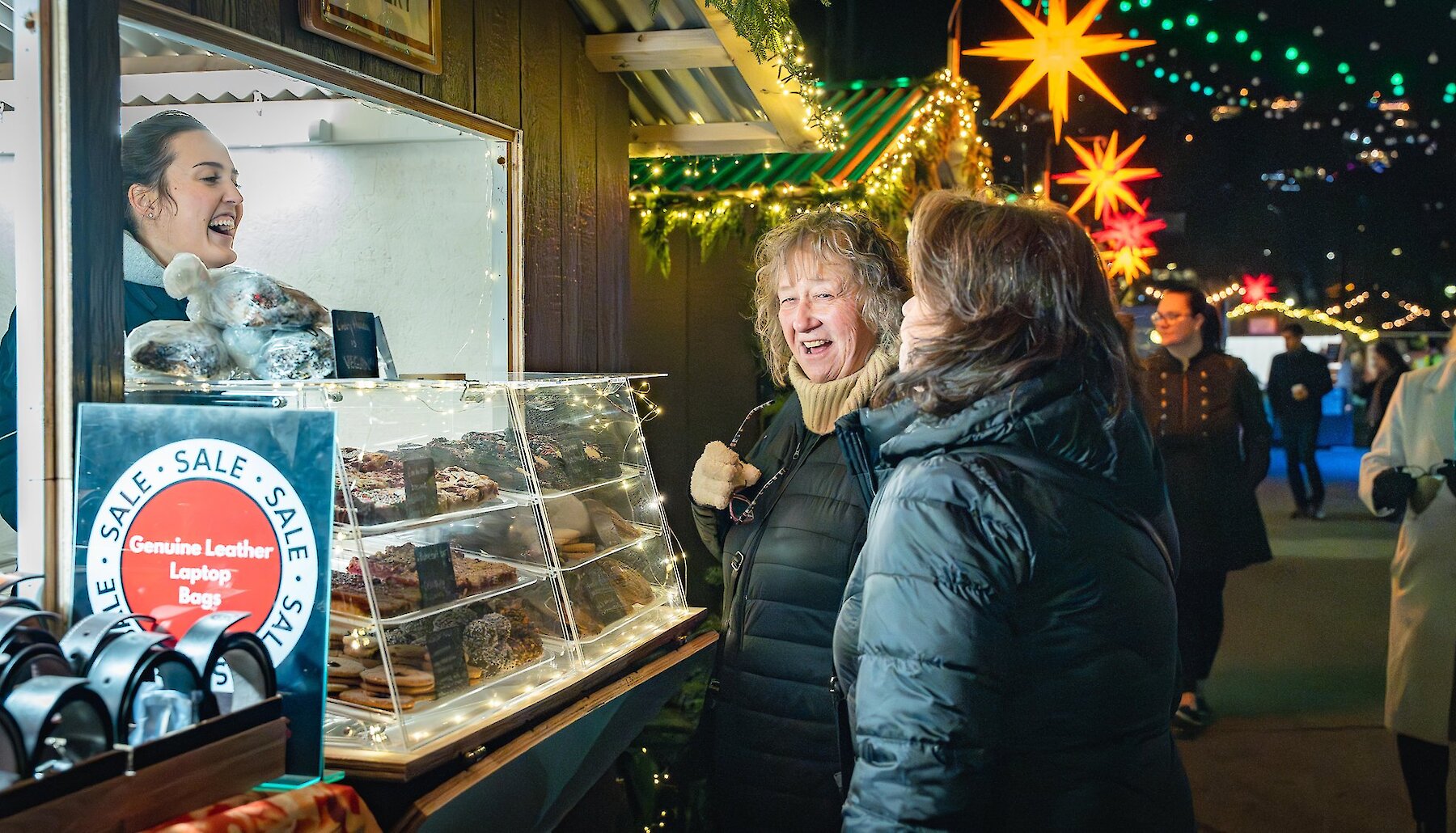 Two shoppers laugh with a vendor at a chalet filled with holiday baking and treats at the Kamloops Christmas Market, with glowing star lanterns overhead.