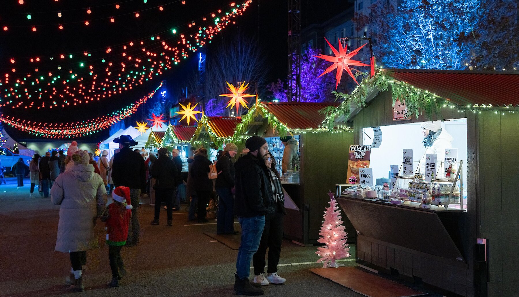 A couple browses handmade fudge, caramel apple cups and hot cocoa jars at the How Sweet! chalet at the Kamloops Christmas Market, under strings of red and green lights and glowing star lanterns.
