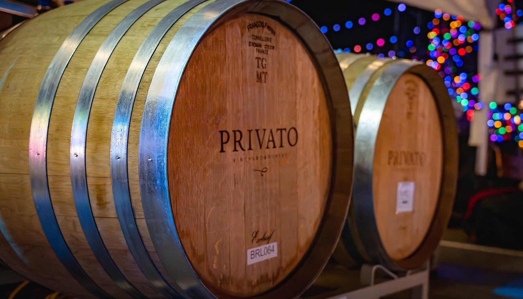 Oak wine barrels branded with the Privato Vineyard & Winery logo, with colourful holiday lights glowing in the background.