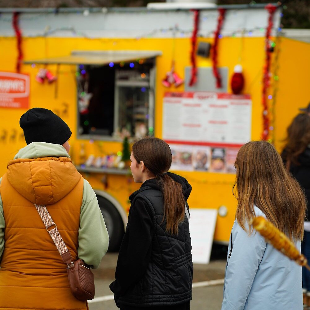 Friends wait in line at the festive yellow Fryers Express food truck, decorated with tinsel and lights, at the Kamloops Christmas Market. One holds a spiral potato on a stick.