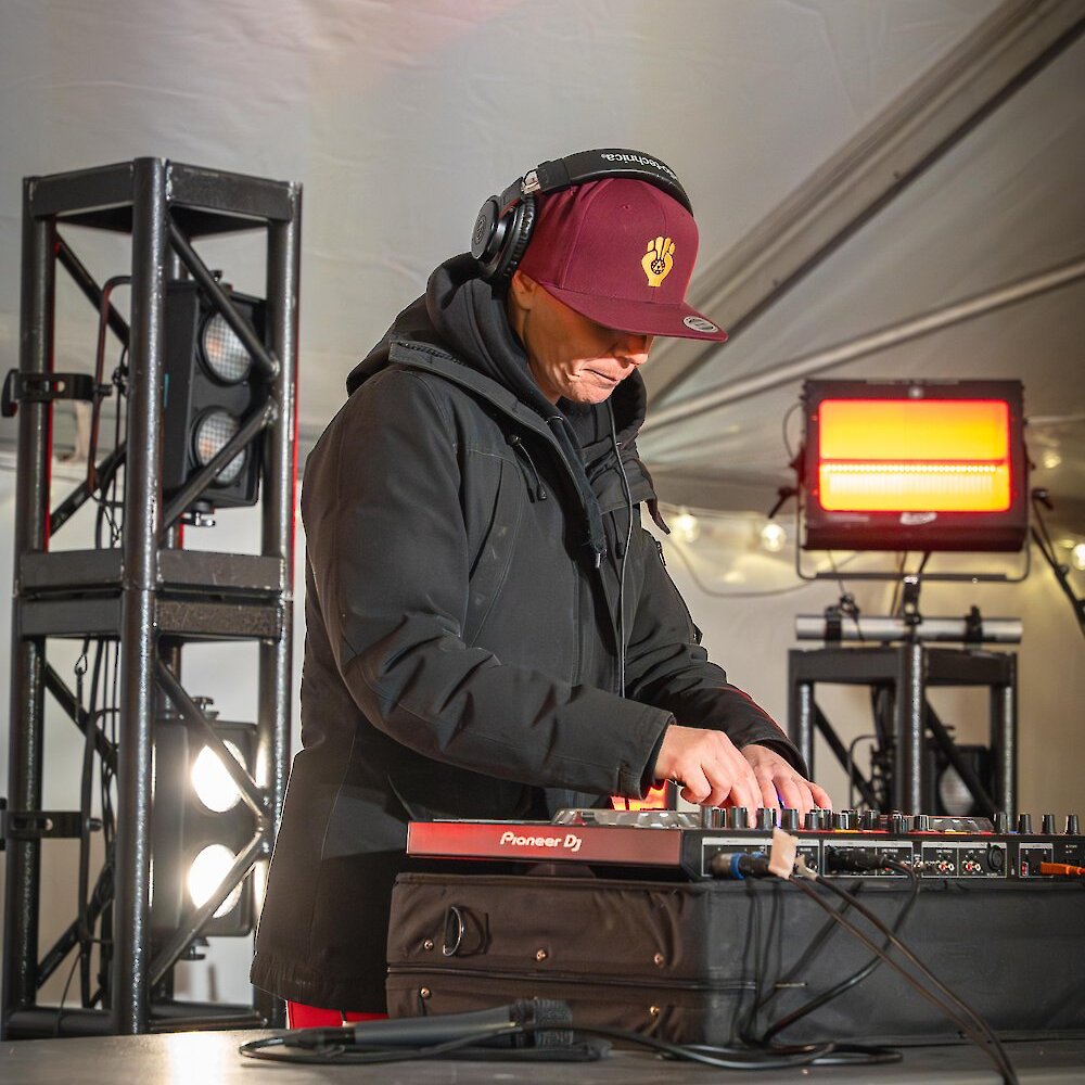 A DJ in headphones and a burgundy cap mixes music on stage under warm lights at the Kamloops Christmas Market.