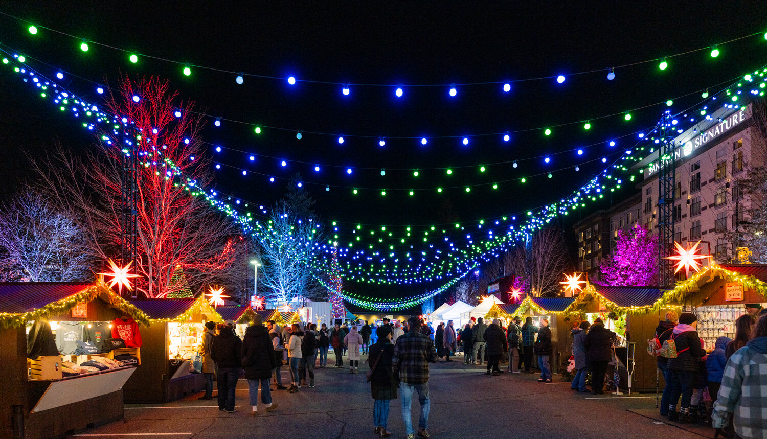 A couple walks arm in arm down the main aisle of the Kamloops Christmas Market at night, under canopies of blue and green string lights between rows of lit wooden chalets.